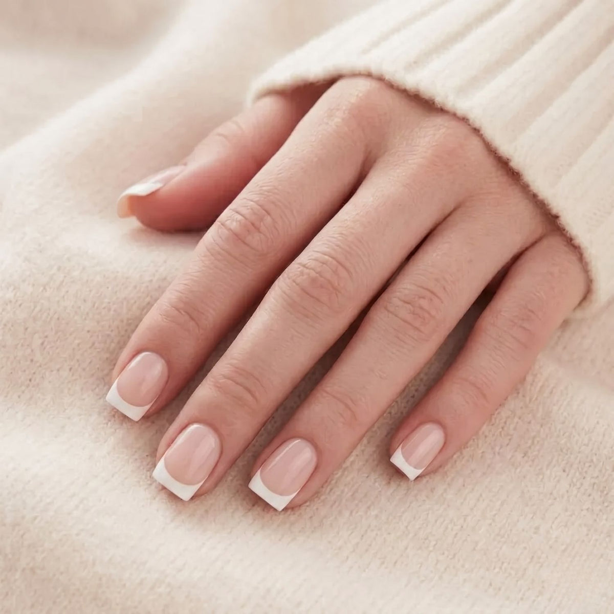 Angelic Aura press-on negle på en hånd mod strikket trøje. Kort, firkantet fransk manicure med nudefarvet bund og hvid spids