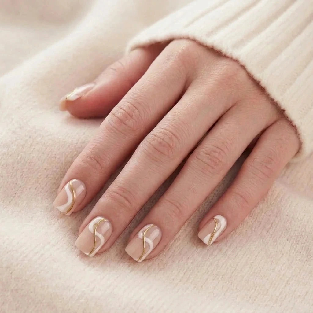 Korte, beige løse negle med hvidt og gyldent bølgedesign på en modelhånd mod en strikbluse.